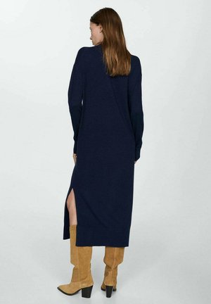 Robe longue à manches longues marine avec fente sur le côté et ourlet côtelé. Tissu en tricot doux ; portée avec des bottes en suède beige jusqu'aux genoux et une silhouette épurée.