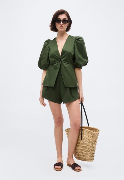 Kvinde iført olivengrøn blazer med puffærmer og matchende shorts, sorte solbriller, sorte slidesandaler, holder stor vævet stråtaske.