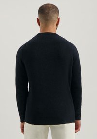Mann mit kurzen Haaren, der einen schwarzen Pullover mit langen Ärmeln und weiße Hosen trägt, steht mit dem Rücken zu einem schlichten Hintergrund.