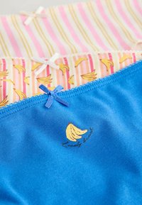Trois paires de sous-vêtements colorés avec des imprimés de bananes et de petits nœuds décoratifs, disposées en couches avec du bleu en haut.