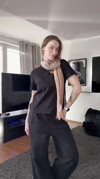 Sort kortærmet top med afslappet pasform, kombineret med sorte bukser med vide ben og et lyst beige tørklæde med diskrete striber. Trægulv og gråt tæppe.