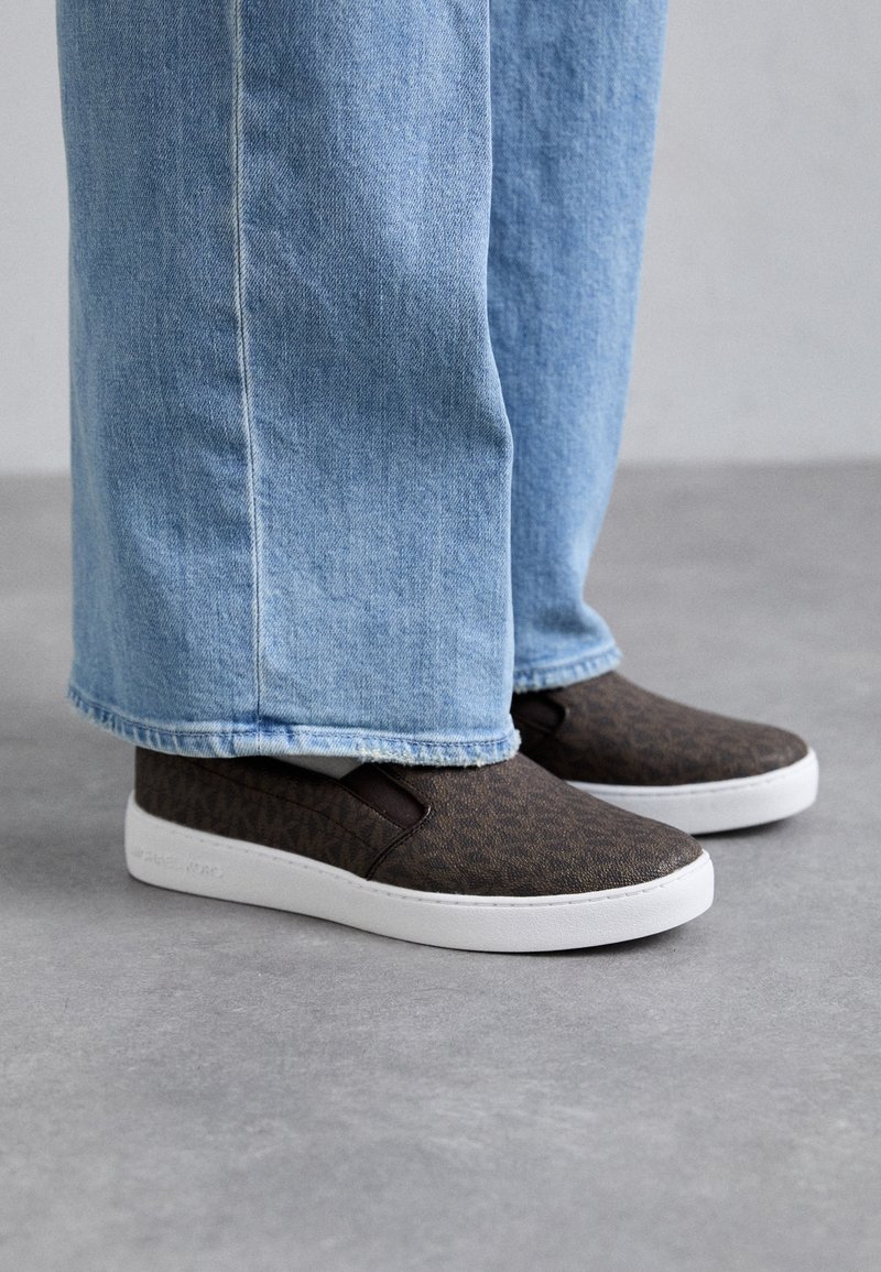 Personne portant un jean large bleu clair et des baskets à enfiler marron foncé avec des semelles blanches, debout sur un sol gris.