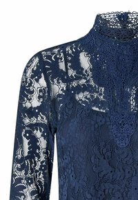 Tmavomodrý čipkovaný top s dlhými rukávmi a vysokým výstrihom, zdobený kvetinovými vzormi a precíznymi detailmi pozdĺž goliera a oblasti na hrudníku.