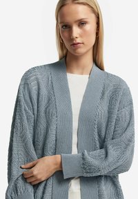 Ljusblå stickad cardigan med texturerade mönster, öppen front och ribbade manschetter. Bärs över en vit topp, med vida ärmar.