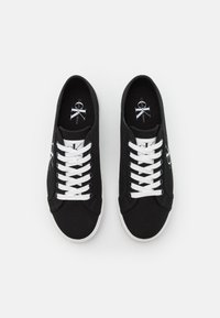 Svarta låga sneakers med vita snören och sulor, med Calvin Klein Jeans-logotyp på tungan och inuti sulan, placerade sida vid sida.