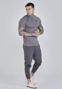 Kratki rukavi, uska siva atletska majica s teksturiranom tkaninom i malim logotipom; u kombinaciji sa odgovarajućim sivim trenirkom i bijelim tenisicama.