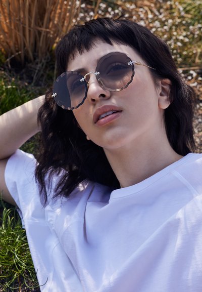 Jeune personne portant de grandes lunettes de soleil rondes festonnées et une chemise blanche, allongée à l'extérieur sur de l'herbe et des plantes séchées au soleil.