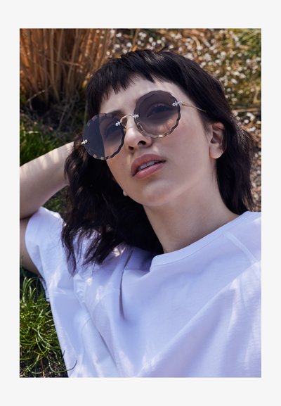 Jeune personne portant de grandes lunettes de soleil rondes festonnées et une chemise blanche, allongée à l'extérieur sur de l'herbe et des plantes séchées au soleil.
