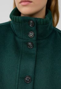 Grüner Wollmantel mit hohem Kragen, vier schwarzen Knöpfen und einem weichen, strukturierten Stoff mit gerippten Details entlang des Kragens.