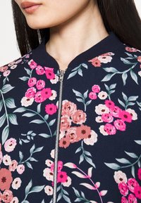 Blommig bomberjacka med en marinblå bas, livliga mönster i rosa och rosor, ribbad krage och en central silverdragkedja.