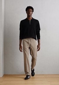 Pull noir à zip quart, pantalon beige et baskets noires avec des rayures blanches. Le modèle se tient devant un fond neutre.