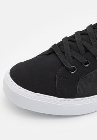 Zapatilla negra con upper de tela suave, panel lateral texturizado y suela de goma blanca. Cuenta con cierre de cordones y ojales redondos.