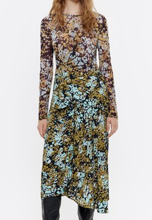 Femme portant une robe à manches longues avec un imprimé floral abstrait aux tons marron, bleu et jaune, associée à des bottes noires montant jusqu'aux genoux.