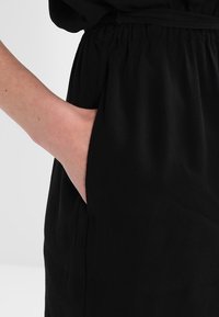 Robe en tissu noir à coupe décontractée, dotée d'une taille élastique et de poches latérales. Texture lisse avec des détails de drapés subtils.