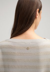 Lichtgrijze trui met horizontale beige strepen, gemaakt van zachte stof. Heeft een ribgebreide halslijn en een ronde logo-accent aan de achterkant.
