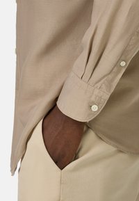 Camisa beige de botones con una textura suave, que presenta un puño enrollado y pequeños botones blancos, combinada con pantalones beige.