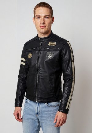 Mann trägt schwarze Lederjacke mit weißen Streifen an den Ärmeln und Aufnähern, kombiniert mit hellblauen Jeans, steht vor einem schlichten Hintergrund.