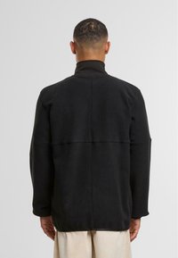 Giacca in fleece nera con colletto alto, dotata di una cucitura orizzontale sulla schiena e maniche lunghe. Morbido al tatto, vestibilità rilassata.