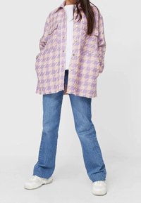 Lila-beige kariertes Shirt-Jacket mit Knopfverschluss, Fronttaschen, lockerer Passform, kombiniert mit blauen Jeans und weißen Sneakers.