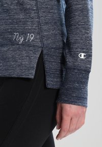 Dettaglio ravvicinato di una persona che indossa una felpa navy blue a trama con "Ny 19" ricamato e il logo Champion sul polsino della manica, abbinata a pantaloni neri.