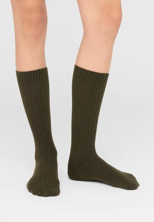 Una modelo lleva calcetines altos hasta la rodilla, de color verde oliva oscuro y acanalados. Se la muestra de las rodillas hacia abajo sobre un fondo blanco.