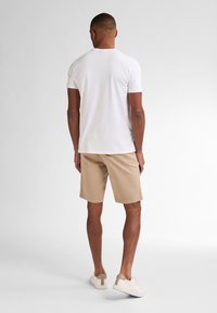 Camiseta blanca de manga corta combinada con pantalones cortos beige; modelo de pie, mostrando líneas limpias y un diseño simple y casual.
