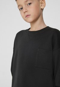 Czarna bluza z miękkiego materiału, z okrągłym dekoltem, luźnym krojem oraz małą, kwadratową kieszenią na piersi. Gładka tekstura.