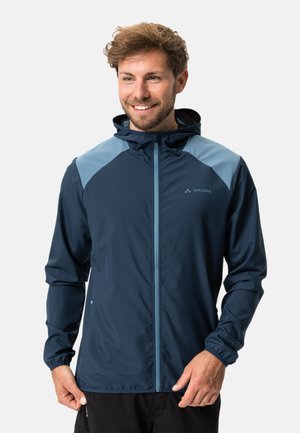 Veste de pluie bleu marine avec des accents bleu clair, fermeture éclair intégrale, capuche ajustable et poignets élastiques. Tissu léger avec une texture lisse.