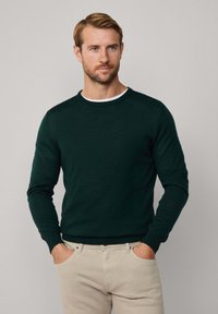 Uomo con capelli castano chiaro e barba, che indossa un maglione a maniche lunghe verde scuro sopra una camicia bianca e pantaloni beige, in piedi con le mani nelle tasche.