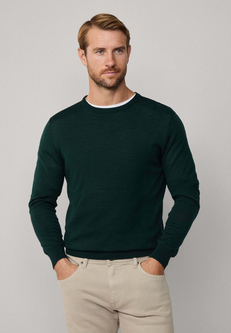 Uomo con capelli castano chiaro e barba, che indossa un maglione a maniche lunghe verde scuro sopra una camicia bianca e pantaloni beige, in piedi con le mani nelle tasche.