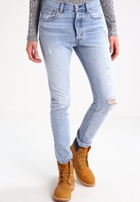 Jeans de denim azul claro desgastados con cortes deshilachados en las rodillas, combinados con botas de encaje color beige. Los jeans presentan un ligero efecto de decoloración y un corte ajustado.