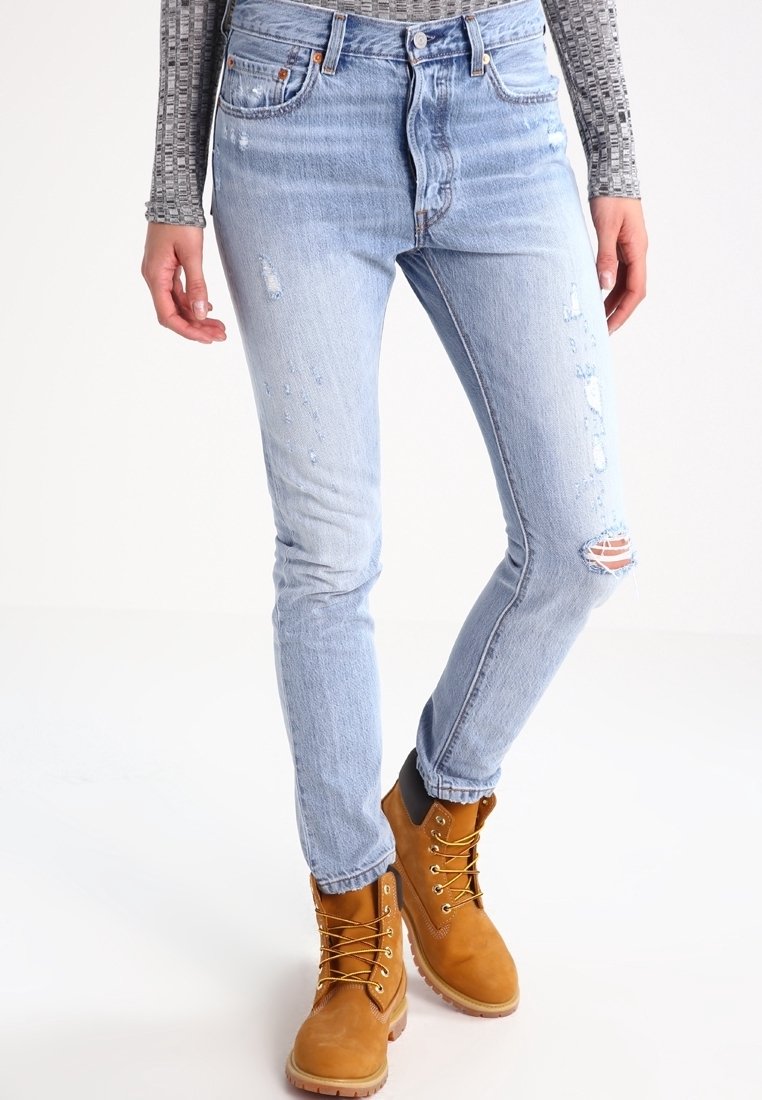 Jeans de denim azul claro desgastados con cortes deshilachados en las rodillas, combinados con botas de encaje color beige. Los jeans presentan un ligero efecto de decoloración y un corte ajustado.