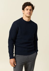 Laivansinis teksturoitu neulepaita pyöreällä kauluksella ja ribbireunuksilla, yhdistettynä harmaisiin housuihin, joissa on vartalonmyötäinen leikkaus.