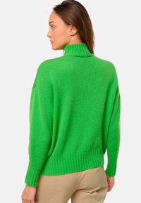 Une femme de profil porte un pull col roulé vert vif et un pantalon beige devant un fond clair et uni.