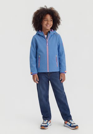 Child with curly hair wearing a blue hooded jacket with pink zippers, dark jeans, and colorful sneakers, standing and smiling.