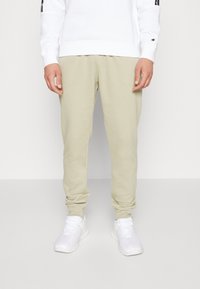 Ljusbeige mjukisbyxor med en avsmalnande design och elastiska muddar, tillverkade av mjukt tyg. Bärs med en vit sweatshirt och vita löparskor.