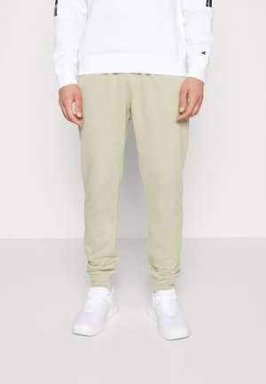 Licht beige sweatpants met een tapered ontwerp en elastische manchetten, gemaakt van zachte stof. Gedragen met een witte sweatshirt en witte sportschoenen.