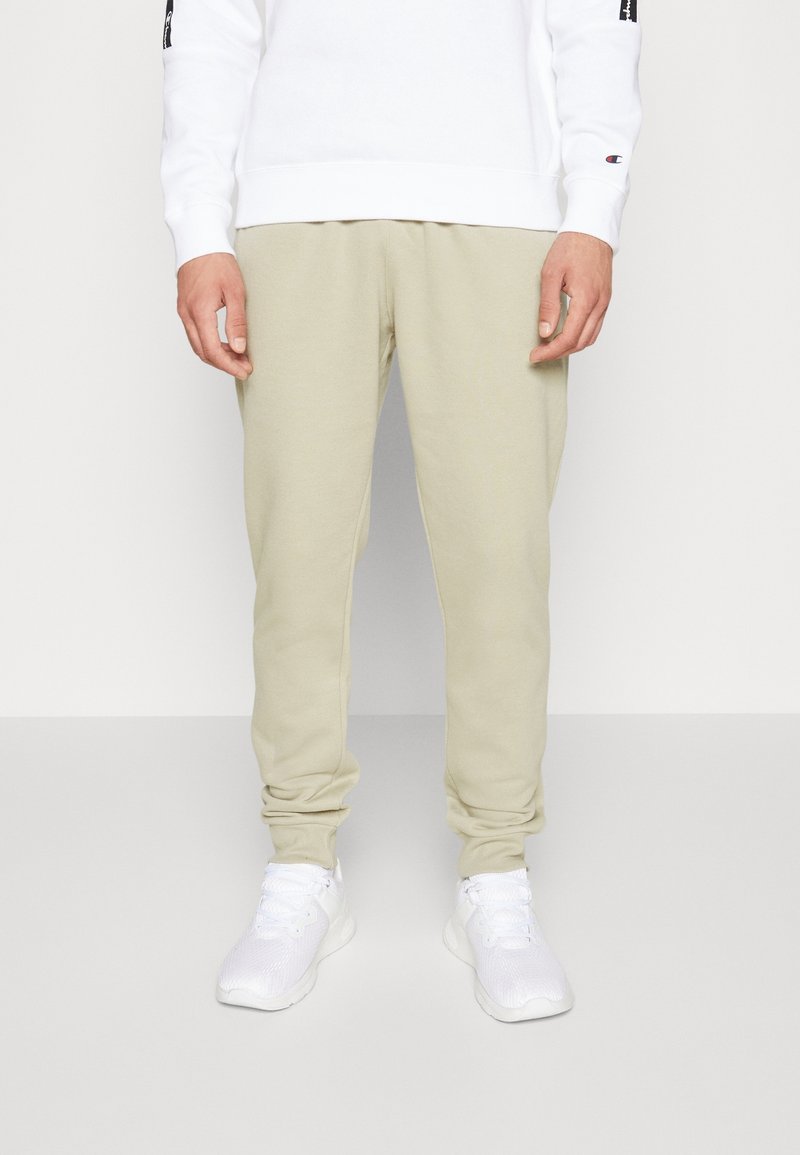 Ljusbeige mjukisbyxor med en avsmalnande design och elastiska muddar, tillverkade av mjukt tyg. Bärs med en vit sweatshirt och vita löparskor.