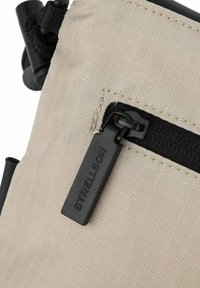 Borsa in tessuto beige con zip nera, dotata di un tirante con marchio. La superficie presenta un motivo a griglia e una texture resistente.