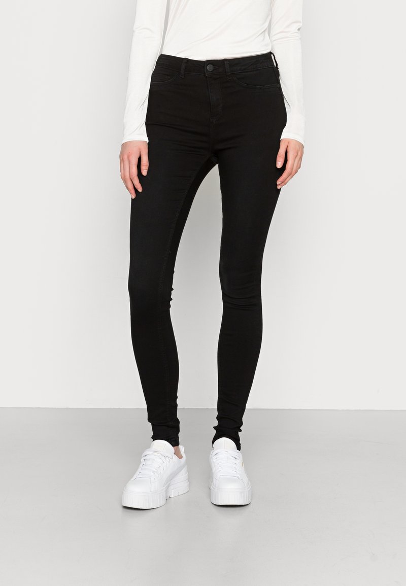 Personne portant un jean skinny noir taille haute et des baskets à plateforme blanches, debout devant un fond uni.