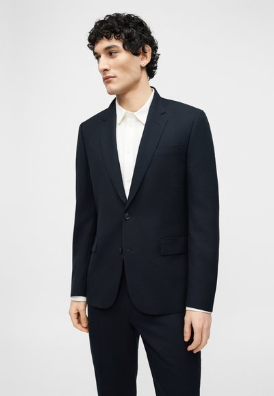 Νεαρός άνδρας με κατσαρά μαλλιά, φορώντας στενό μαύρο κοστούμι και λευκό πουκάμισο με κουμπιά, κοιτάζει προς τα αριστερά σε ένα απλό ανοιχτό υπόβαθρο.