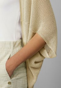 Goldfarbige Netzstrickjacke mit kurzen Ärmeln, über einem weißen Top und beigen Textilhosen getragen, verfügt über eine Tasche und goldene Knopfdetails.