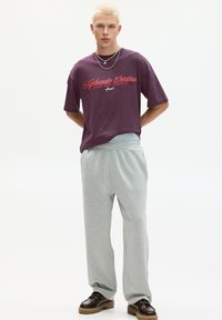 Lilla grafisk t-shirt med rød tekst, kombineret med lysegrå sweatpants. Modellen bærer mørke sko og lagdelte sølvhalskæder.