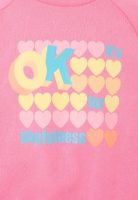 Sweat à capuche rose avec un grand texte "OK" en bleu et jaune, entouré de cœurs de différentes couleurs pastel, accompagné de la phrase "c'est pour le côté ludique."