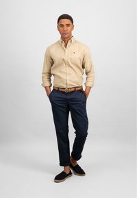 Beige linnen overhemd met knopen, marineblauwe chino's, bruine gevlochten riem, zwarte espadrilles met jute zolen. Model staat in een neutrale omgeving.