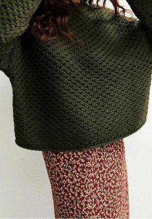 Femme portant un pull en grosse maille texturé vert foncé et une jupe fleurie rouge taille haute, les bras levés au-dessus de la tête.