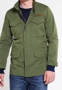Olivgrön jacka gjord av tyg, med fyra framfickor, en ståkrage och dragkedjestängning. Ytan är slät med en matt finish.