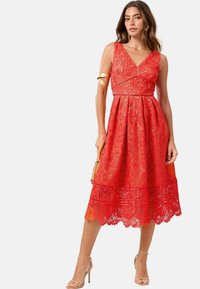 Rotes Spitzenkleid mit V-Ausschnitt und ärmellosem Design. Detailliertes Blumendesign, ausgestellter Rock und gewellter Saum. Kombiniert mit nude-farbenen High Heels.