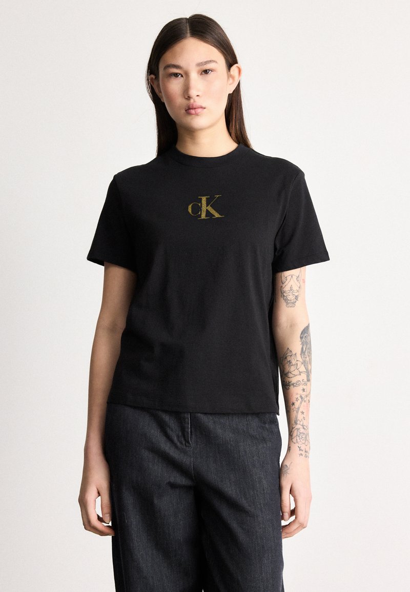 Jeune femme aux longs cheveux foncés portant un T-shirt noir Calvin Klein et un pantalon sombre, montrant des tatouages sur son avant-bras gauche.