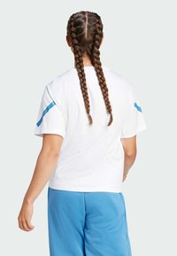 Camiseta blanca de manga corta con un acento azul en el hombro, complementada con pantalones cortos deportivos azules. Se puede ver el cabello trenzado por la espalda.